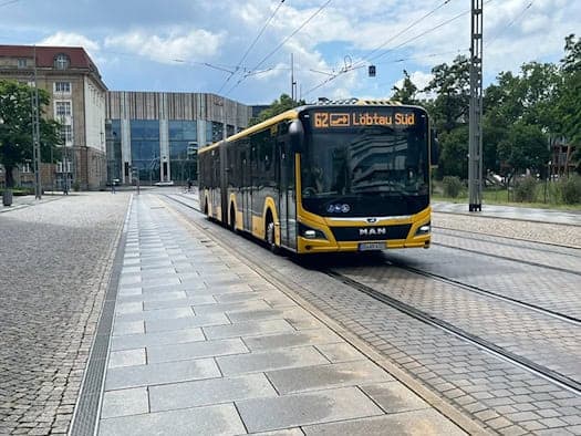Ein gelb-schwarzer Gelenkbus der DVB mit der Anzeige „62 Löbtau Süd“ fährt auf einer gepflasterten Straße mit Schienen. Im Hintergrund sind moderne und historische Gebäude unter leicht bewölktem Himmel zu sehen.