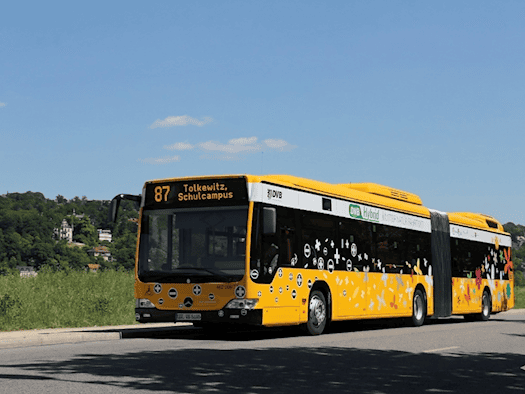 Ein gelb-schwarzer Hybrid-Gelenkbus der Dresdner Verkehrsbetriebe mit der ki-generierten, digitalen Zielanzeige "87 Tolkewitz, Schulcampus" fährt auf einer Straße in Dresden.