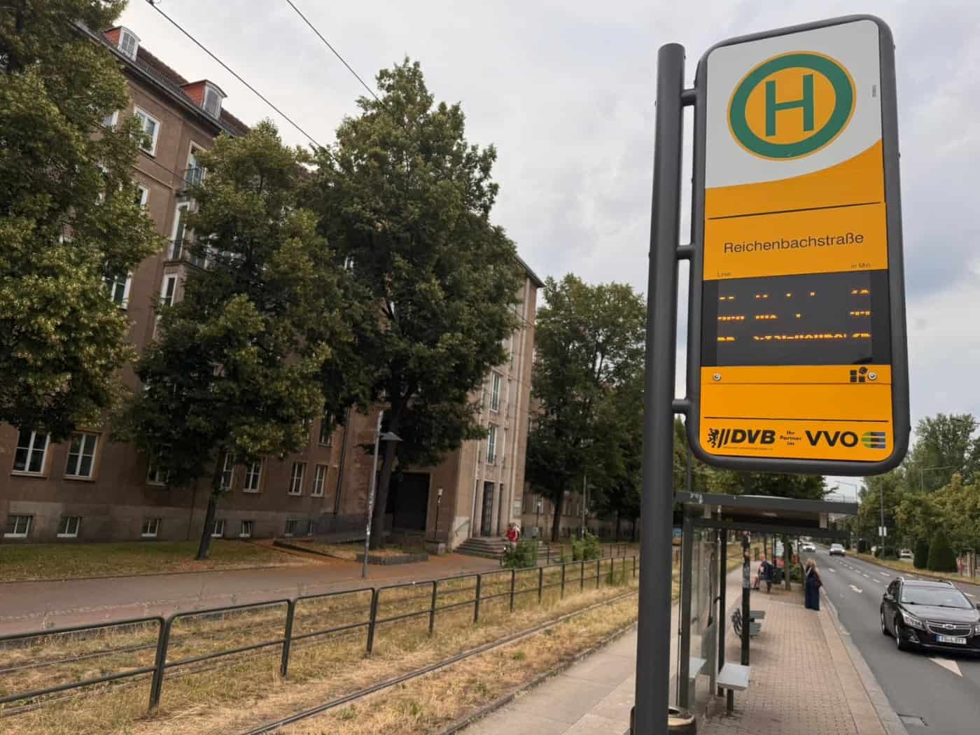 Nahaufnahme des gelb-grauen Haltestellenschildes Reichenbachstraße der DVB mit einer digitalen Anzeige. Im Hintergrund sind Wohngebäude und grüne Bäume an der Fritz-Löffler-Straße unter wolkenverhangenem Himmel zu sehen.