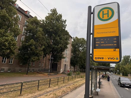 Nahaufnahme des gelb-grauen Haltestellenschildes Reichenbachstraße der DVB mit einer digitalen Anzeige. Im Hintergrund sind Wohngebäude und grüne Bäume an der Fritz-Löffler-Straße unter wolkenverhangenem Himmel zu sehen.