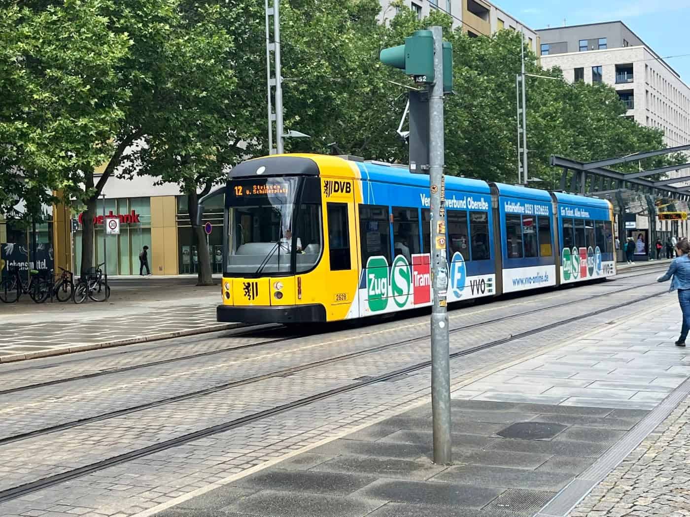 Eine moderne, gelb-blaue Straßenbahn der Linie 12 fährt auf Schienen an einer Allee mit grünen Bäumen und hellen Wohngebäuden vorbei. Im Vordergrund steht ein grauer Ampelmast.