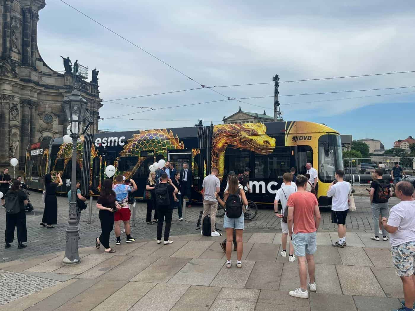 Eine moderne, schwarz folierte Straßenbahn mit einem großen, goldenen Drachenkopf an der Seite steht an einem Platz vor einem historischen Sandsteingebäude. Passanten stehen davor und fotografieren das Fahrzeug.