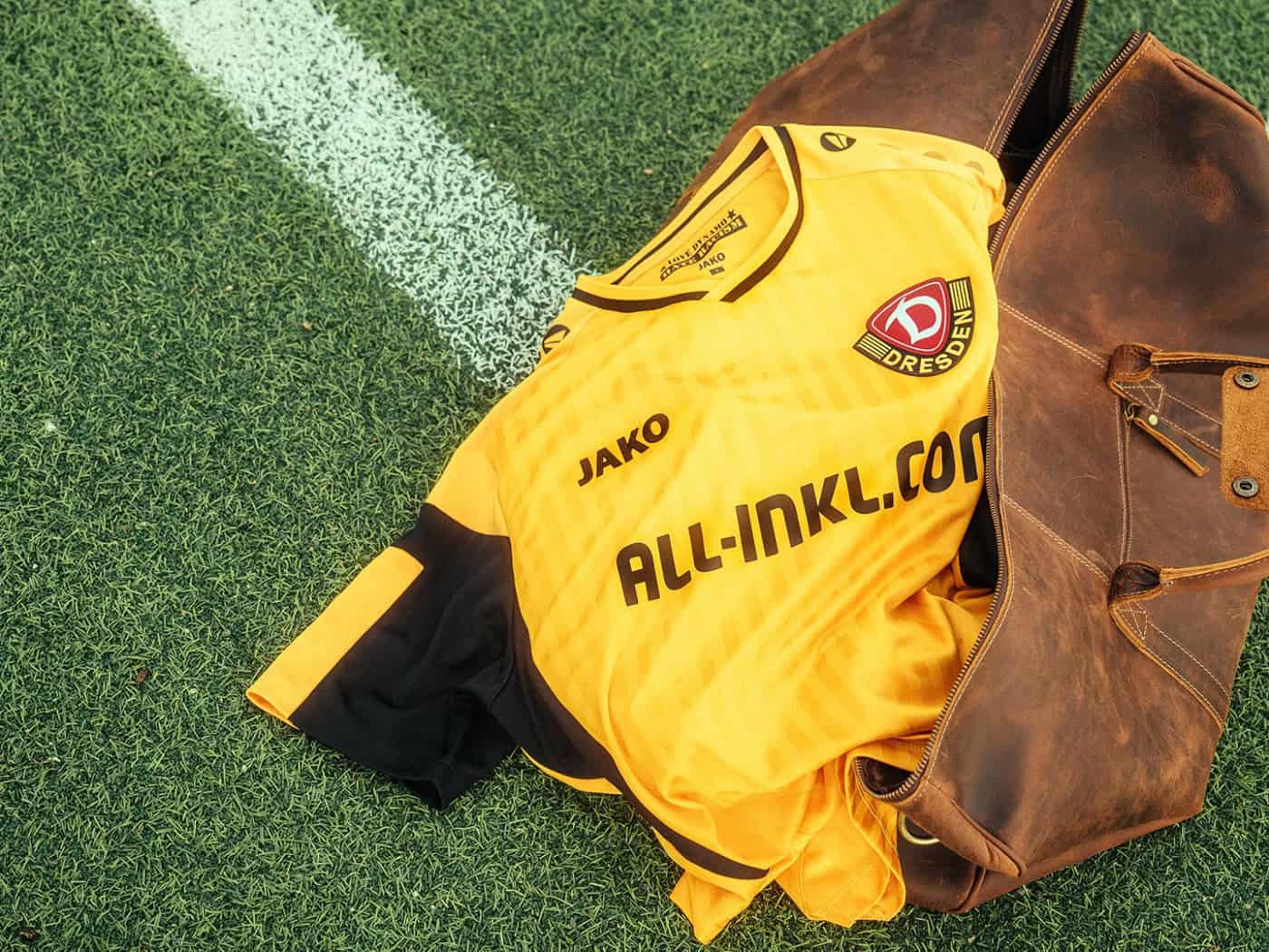 Ein gelb-schwarzes Dynamo-Dresden-Trikot liegt in einer offenen, braunen Ledertasche auf grünem Kunstrasen direkt neben einer weißen Spielfeldmarkierung.