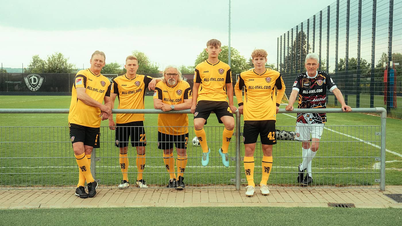 Sechs Männer, bestehend aus älteren Vereinslegenden und jungen Fußballern in gelb-schwarzen Trikots, stehen nebeneinander an einem Zaun auf einem Sportplatz.
