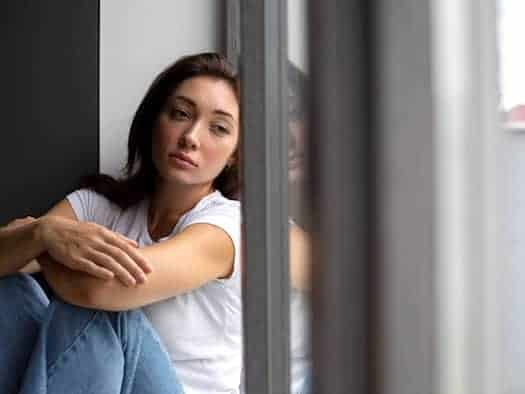 Eine junge Frau im weißen T-Shirt und Jeans sitzt mit angezogenen Beinen traurig am Boden neben einer großen Fensterscheibe. Ihr ernster Gesichtsausdruck spiegelt sich im Glas, während ihr Blick melancholisch ins Leere geht.
