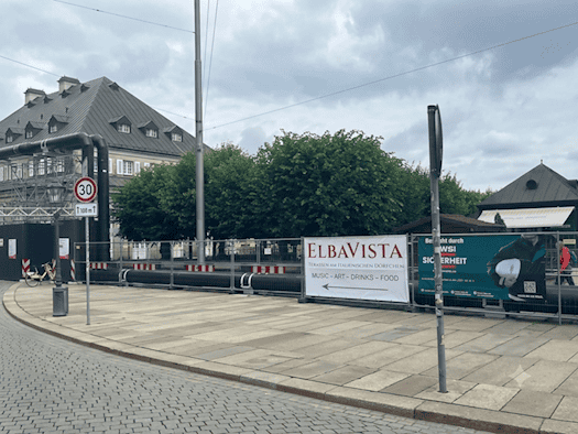 Ein großes weißes Werbebanner mit der Aufschrift „ElbaVista Terrassen am Italienischen Dörfchen – Music – Art – Drinks – Food“ hängt an einem Bauzaun neben einem gepflasterten Gehweg. Im Hintergrund ist ein historisches Gebäude zu sehen, links verlaufen große schwarze Rohrleitungen.