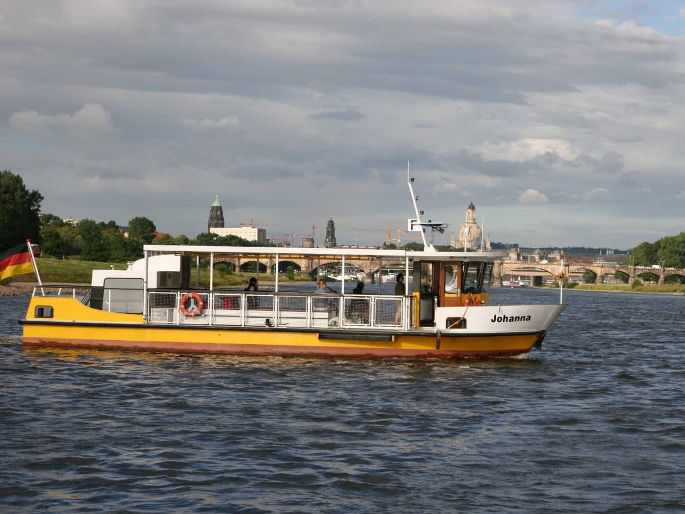 Das gelb-weiße Fährboot Johanna der Dresdner Verkehrsbetriebe fährt auf der Elbe vor der Kulisse der Dresdner Altstadt mit Frauenkirche und Augustusbrücke.