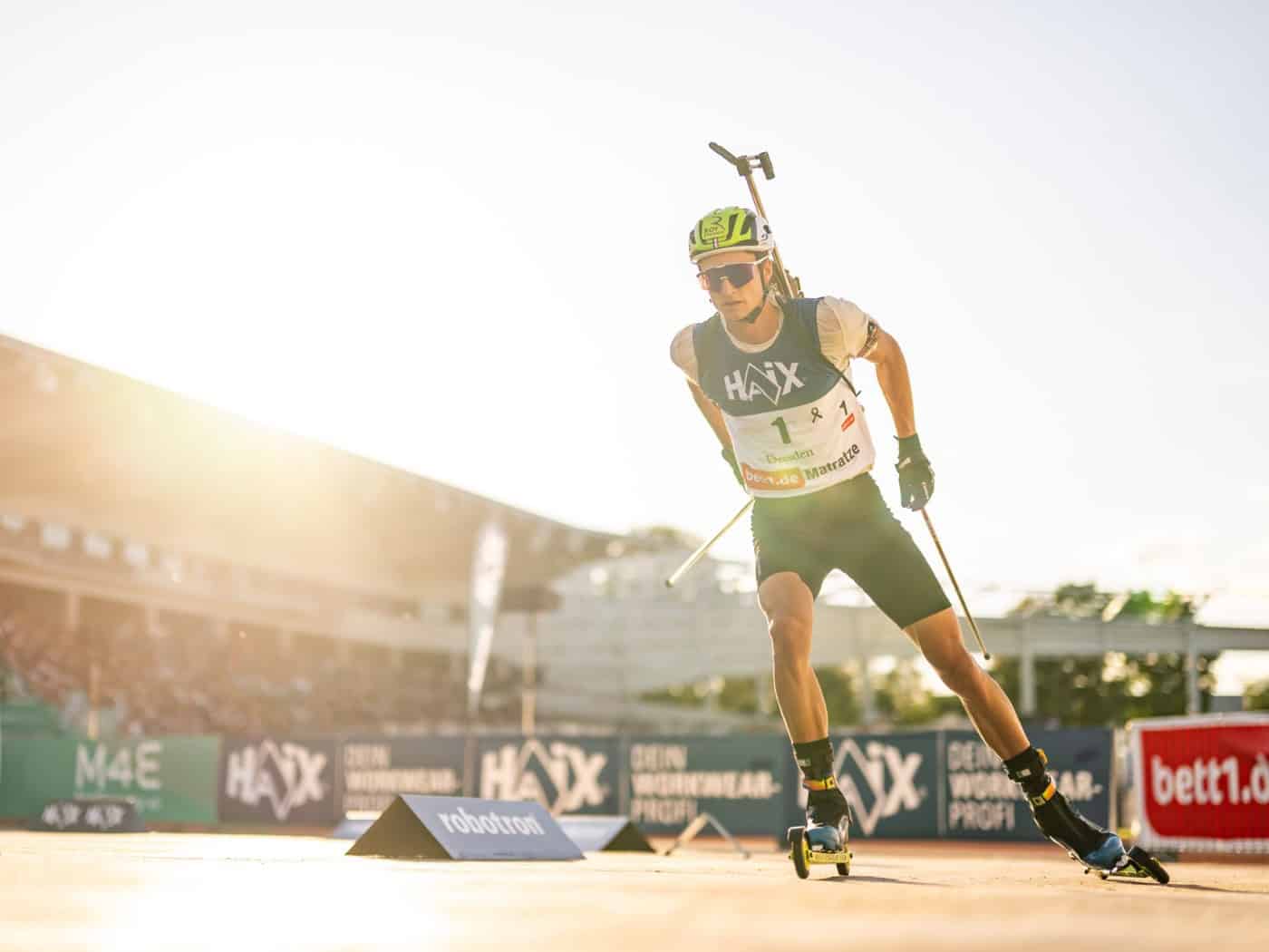 Ein männlicher Biathlet läuft auf Skirollern mit hoher Geschwindigkeit auf der Zielgeraden im Heinz-Steyer-Stadion in Dresden. Er trägt ein weißes Renntrikot mit der Startnummer 1 und dem Sponsoren-Schriftzug HAIX, eine dunkle Sport-Radlerhose, eine Sportsonnenbrille und einen neongelben Schutzhelm.