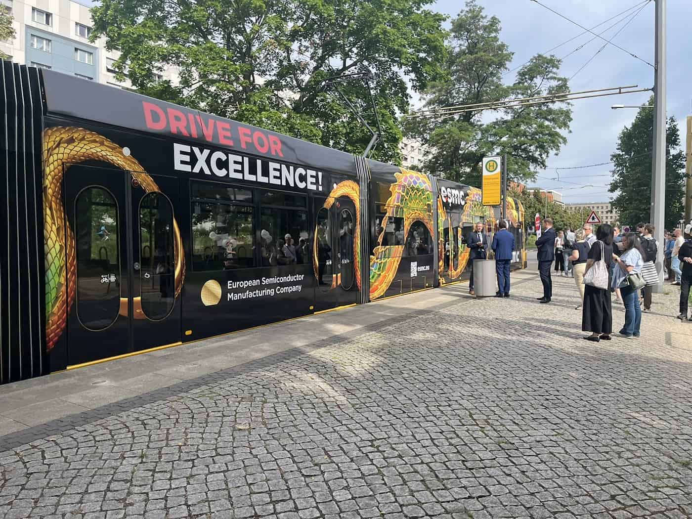 A black-and-yellow tram is parked at a cobblestone stop. The words “DRIVE FOR EXCELLENCE!” are emblazoned on the side of the vehicle against a golden dragon-scale pattern. Several people are standing on the sidewalk next to it, waiting.