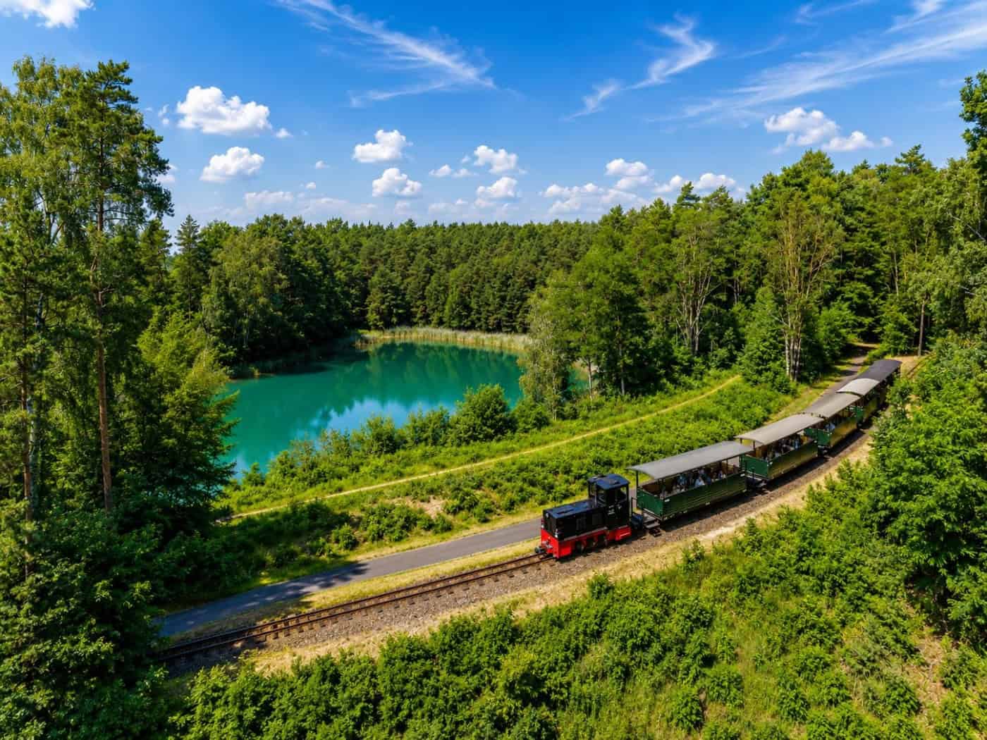 Eine kleine Schmalspur-Diesellok zieht mehrere offene Personenwagen durch eine waldige Landschaft, im Hintergrund ein kleiner türkisfarbener See