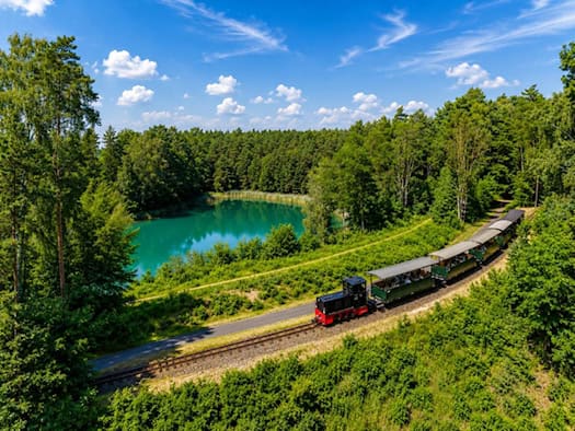 Eine kleine Schmalspur-Diesellok zieht mehrere offene Personenwagen durch eine waldige Landschaft, im Hintergrund ein kleiner türkisfarbener See