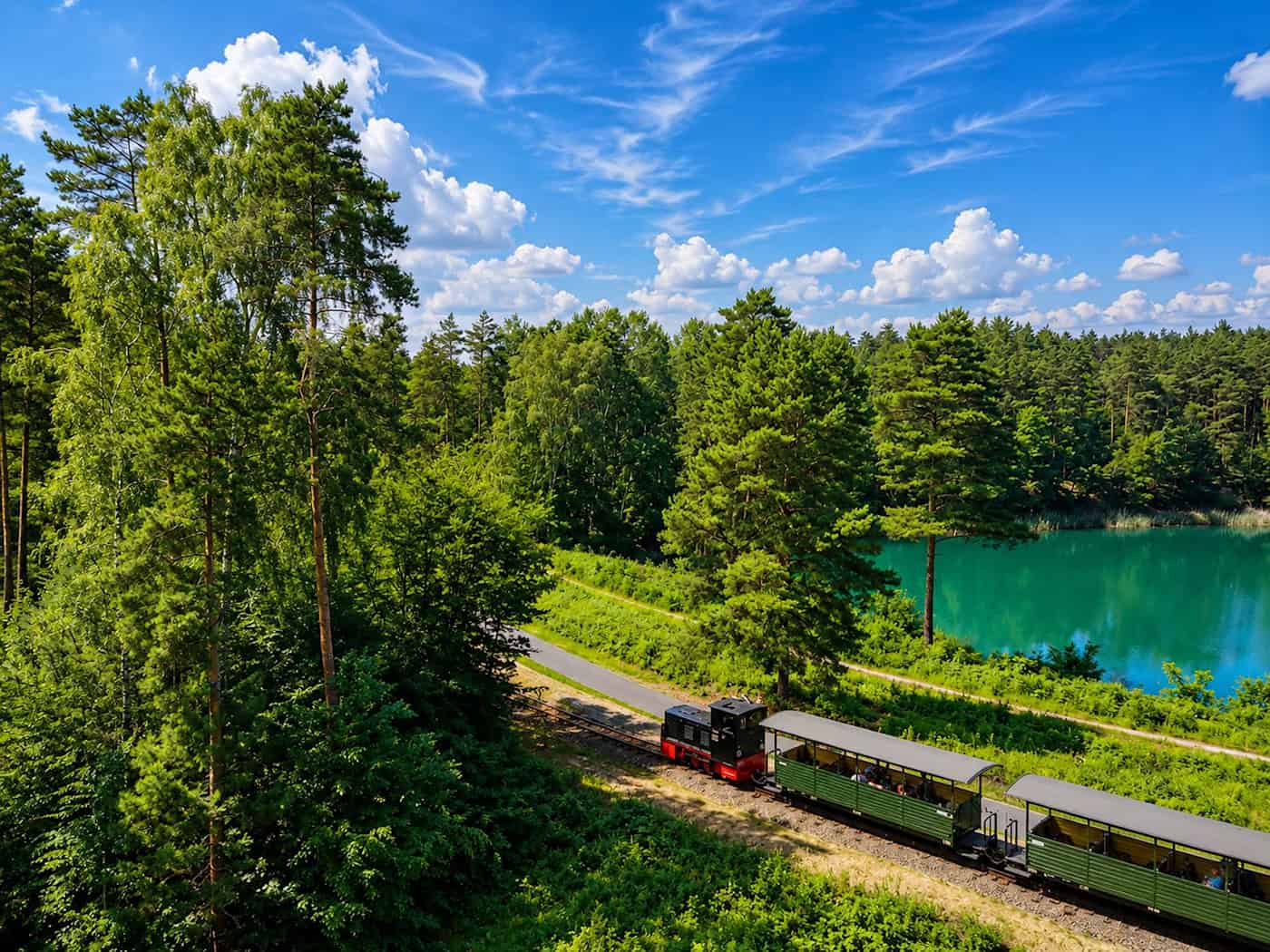 Eine kleine Schmalspur-Diesellok zieht mehrere offene Personenwagen durch eine waldige Landschaft, im Hintergrund ein kleiner türkisfarbener See