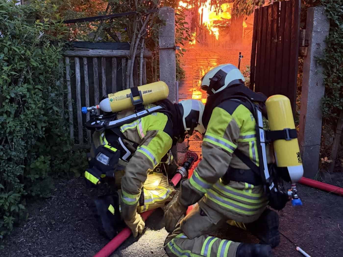 Zwei Feuerwehrleute in reflektierender Schutzkleidung mit gelben Atemschutzflaschen knien im Vordergrund an einem roten Löschschlauch. Durch ein hölzernes Gartentor im Hintergrund sind hell lodernde, orangefarbene Flammen zu sehen.