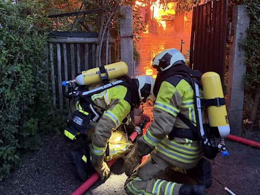 Zwei Feuerwehrleute in reflektierender Schutzkleidung mit gelben Atemschutzflaschen knien im Vordergrund an einem roten Löschschlauch. Durch ein hölzernes Gartentor im Hintergrund sind hell lodernde, orangefarbene Flammen zu sehen.