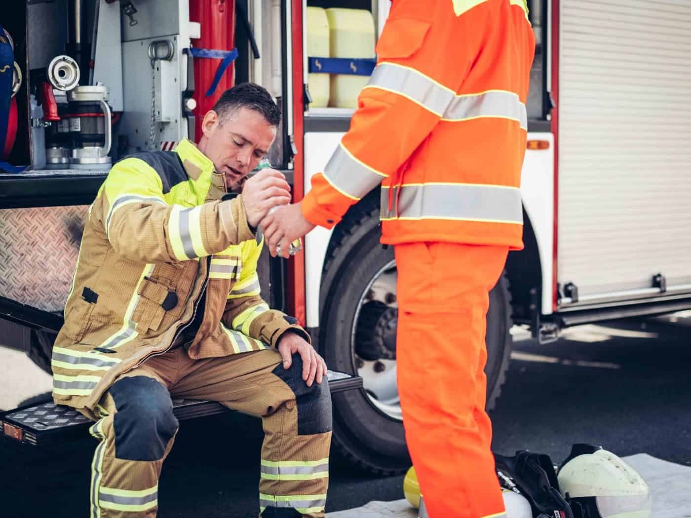 Ein erschöpfter Feuerwehrmann sitzt in voller Schutzbekleidung auf dem Trittbrett eines Feuerwehrfahrzeugs und nimmt eine Wasserflasche entgegen.