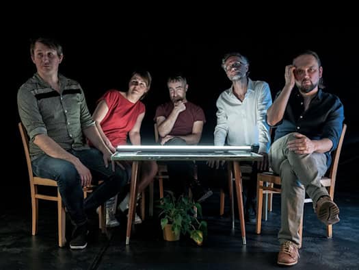 Die fünf Bandmitglieder von Fiffy & The Kids sitzen im schummrigen Licht um einen von unten beleuchteten Tisch vor schwarzem Hintergrund.