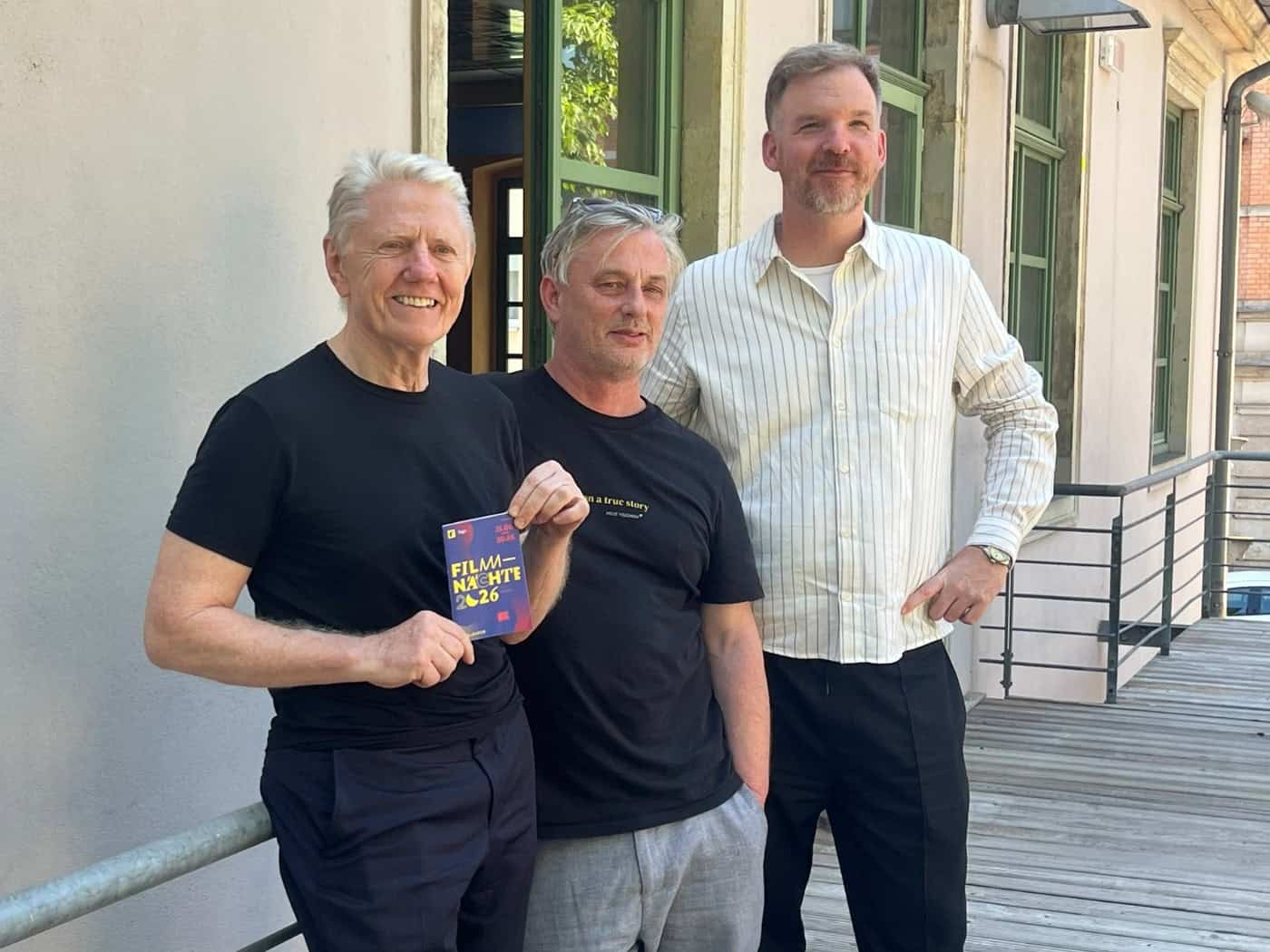 Drei lächelnde Männer auf der Holzterrasse des ehemaligen Astoria-Kinos in Dresden. Der linke Mann mit weißen Haaren hält einen blauen Flyer mit der Aufschrift „Filmnächte 2026“ in der Hand.