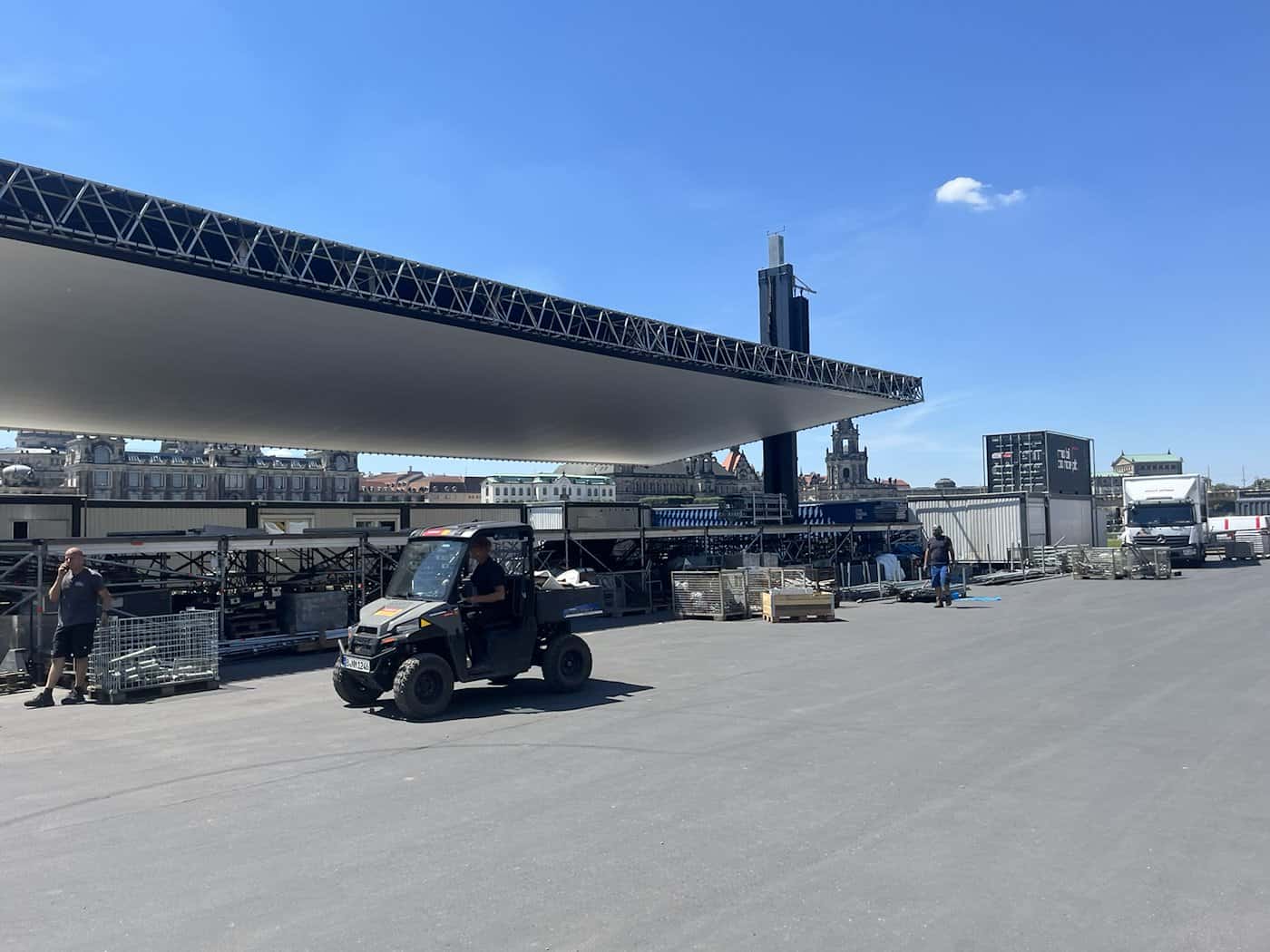 Großbaustelle der Filmnächte am Elbufer in Dresden bei strahlendem Sonnenschein. Eine riesige, flach montierte Kinoleinwand auf Gerüstteilen dominiert das asphaltierte Gelände, auf dem ein kleines Nutzfahrzeug fährt. Im Hintergrund ist die Silhouette der Dresdner Altstadt zu sehen.