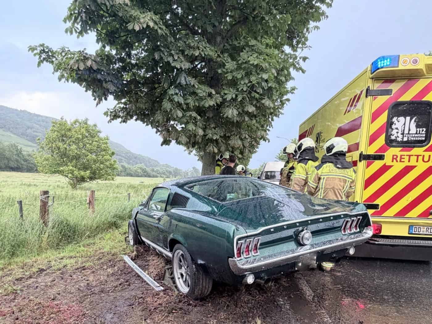 Die Heck- und Seitenansicht eines dunkelgrünen Ford Mustang Oldtimers, der auf dem unbefestigten, schlammigen Randstreifen direkt vor einem großen, belaubten Laubbaum steht. Rechts neben dem Unfallwagen parkt ein gelb-roter Rettungswagen der Feuerwehr Dresden mit eingeschaltetem Blaulicht.