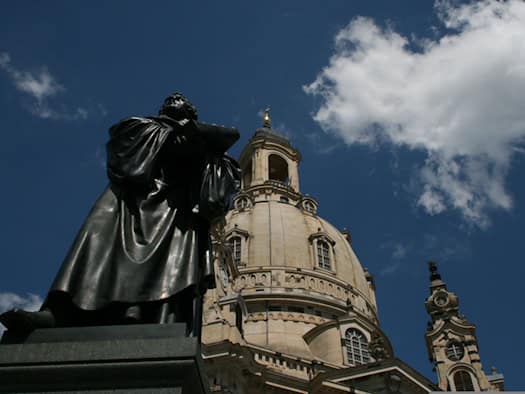 Froschperspektive auf das dunkle Martin-Luther-Standbild aus Bronze vor der hellen Steinkuppel der Dresdner Frauenkirche unter einem blauen Himmel mit weißen Wolken. 