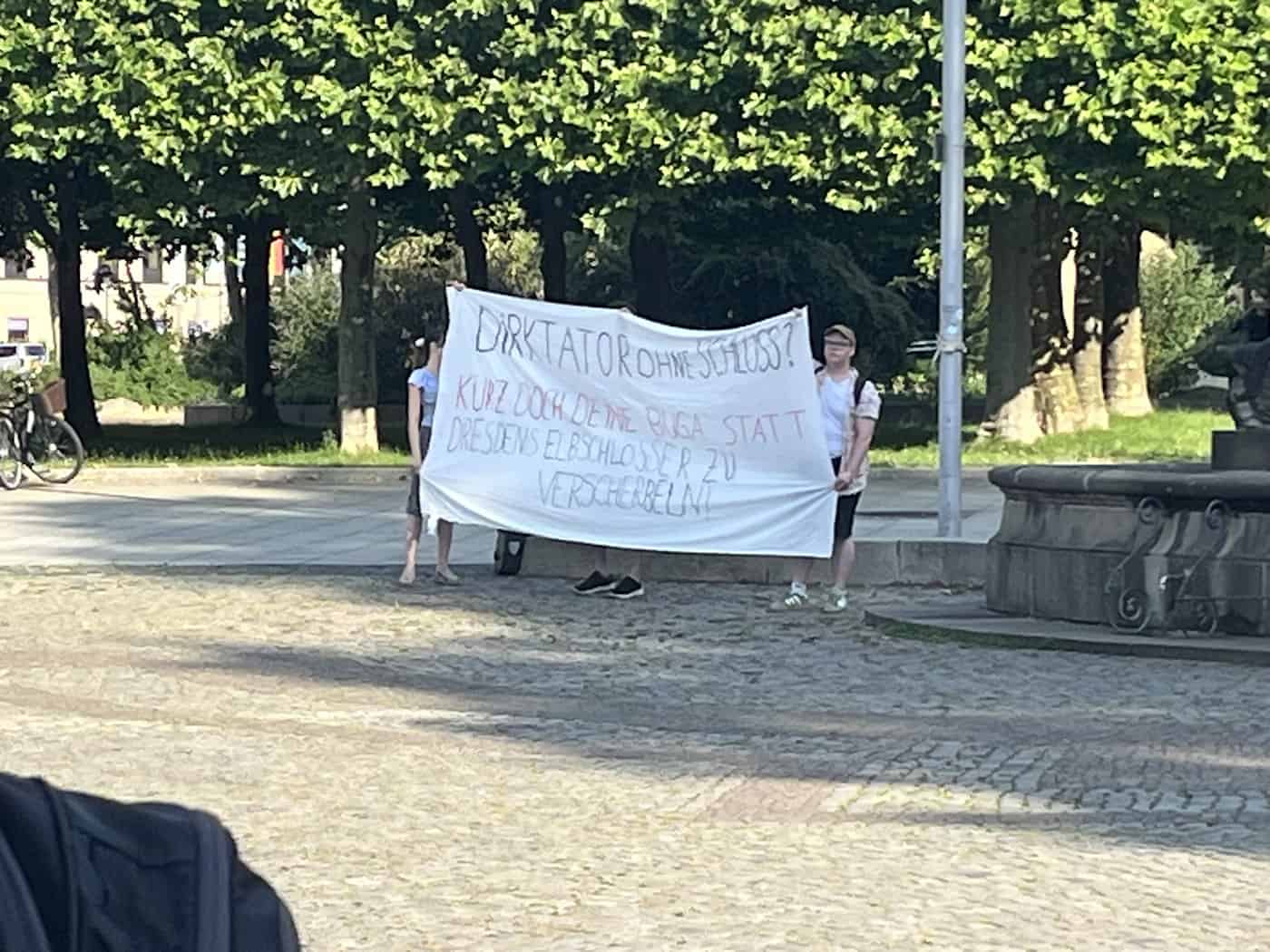 Drei Personen halten auf einem gepflasterten Platz ein großes, weißes, handgemaltes Transparent mit der Aufschrift „Dirktator ohne Schloss? Kürz doch deine BUGA, statt Dresdens Elbschlösser zu verscherbeln!“.