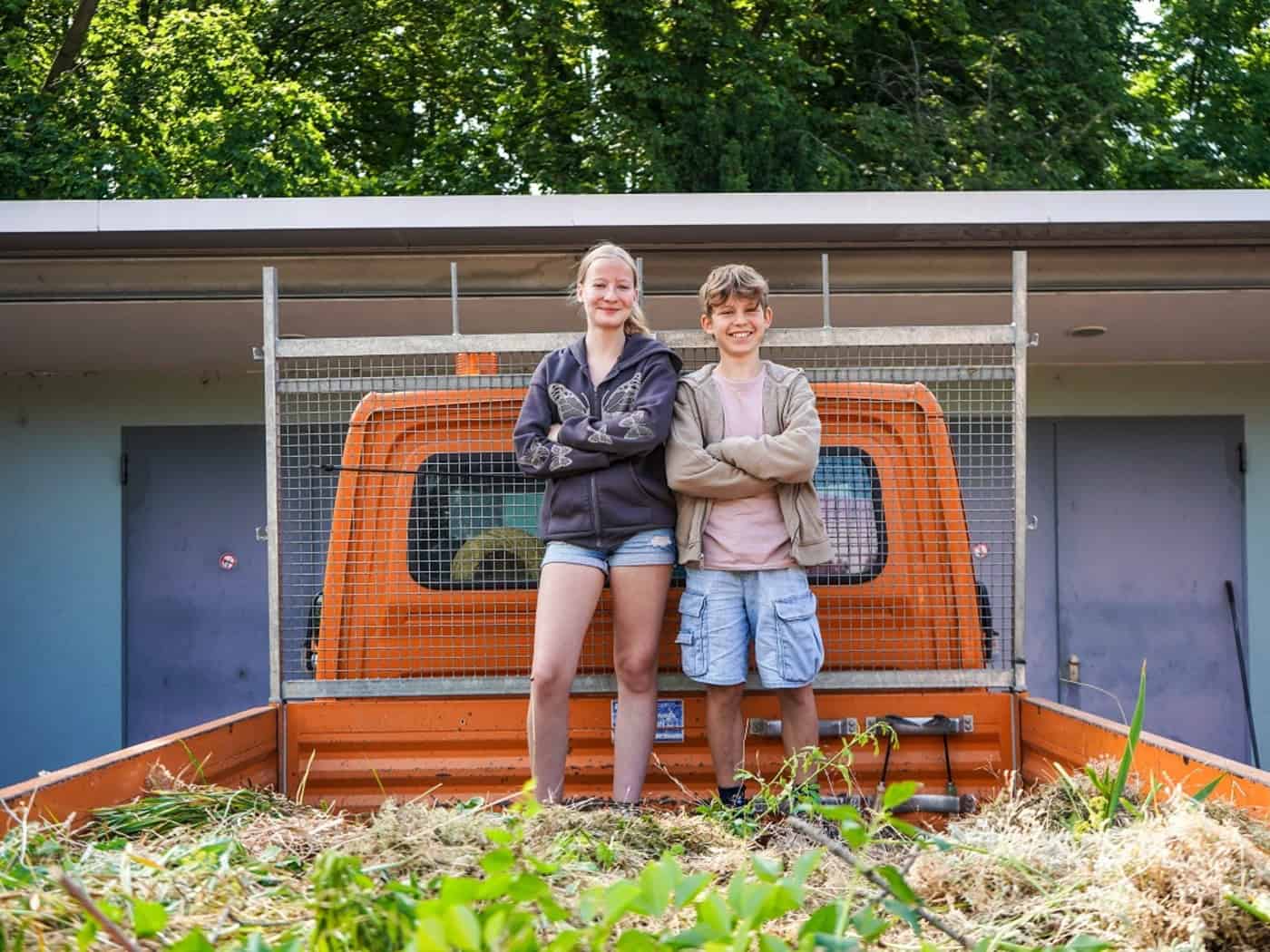 Ein lächelndes Mädchen und ein Junge stehen mit verschränkten Armen auf der Ladefläche eines orangefarbenen Lkw, die mit abgeschnittenem Gras und Zweigen gefüllt ist. Im Hintergrund sind grüne Bäume zu sehen.