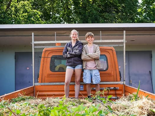 Ein lächelndes Mädchen und ein Junge stehen mit verschränkten Armen auf der Ladefläche eines orangefarbenen Lkw, die mit abgeschnittenem Gras und Zweigen gefüllt ist. Im Hintergrund sind grüne Bäume zu sehen.