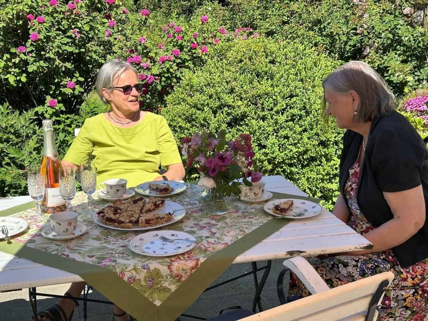 zwei ältere Damen sitzen im Grünen bei Kuchen und Sekt an einem Tisch und unterhalten sich