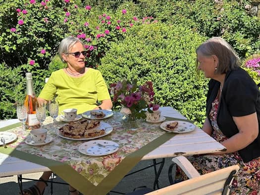 zwei ältere Damen sitzen im Grünen bei Kuchen und Sekt an einem Tisch und unterhalten sich