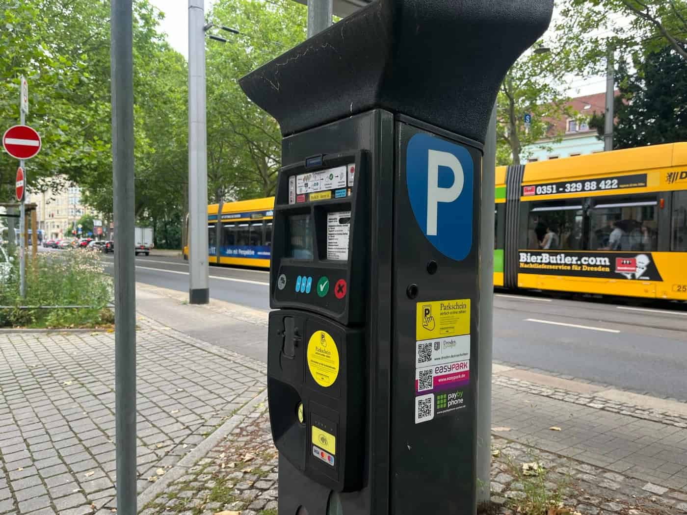 Ein grauer Parkscheinautomat am Straßenrand in Dresden mit bunten Aufklebern für mobiles Parken wie EasyPark. Im Hintergrund fährt eine gelbe Straßenbahn der DVB vor grünen Bäumen vorbei.