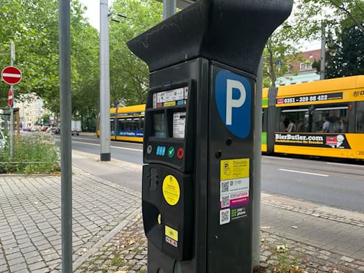 Ein grauer Parkscheinautomat am Straßenrand in Dresden mit bunten Aufklebern für mobiles Parken wie EasyPark. Im Hintergrund fährt eine gelbe Straßenbahn der DVB vor grünen Bäumen vorbei.