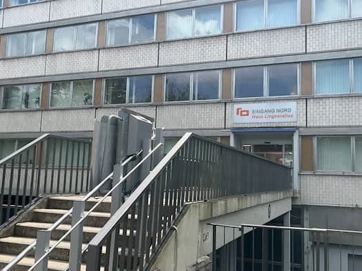Außenansicht eines mehrstöckigen Bürogebäudes mit grauer Fliesenstruktur. Eine Betonrampe mit grauem Metallgeländer und einem montierten Plattformlift für Rollstühle führt zum Eingang mit dem Schild „EINGANG NORD Haus Lingnerallee“.