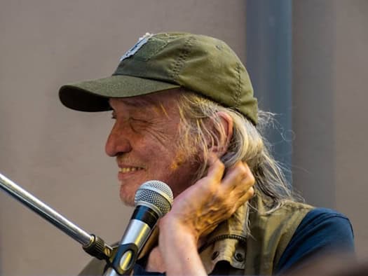 Foto: Lächelnder Mann mit längerem grauen Haar und grüner Basecap im Profil. Vor ihm hängt ein Mikrofon, linke Hand am Hals.