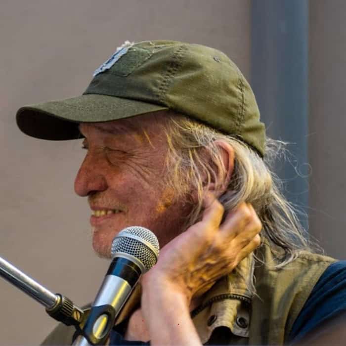 Foto: Lächelnder Mann mit längerem grauen Haar und grüner Basecap im Profil. Vor ihm hängt ein Mikrofon, linke Hand am Hals.