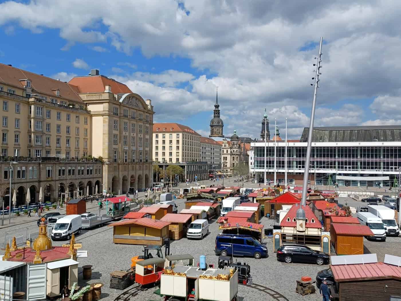 Blick von oben auf den Dresdner Altmarkt mit hölzernen Markthütten und weißen Transportern. Im Hintergrund stehen mehrstöckige Gebäude und die Glasfront des Kulturpalasts unter blauem Himmel.
