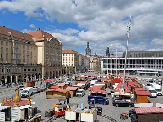 Blick von oben auf den Dresdner Altmarkt mit hölzernen Markthütten und weißen Transportern. Im Hintergrund stehen mehrstöckige Gebäude und die Glasfront des Kulturpalasts unter blauem Himmel.