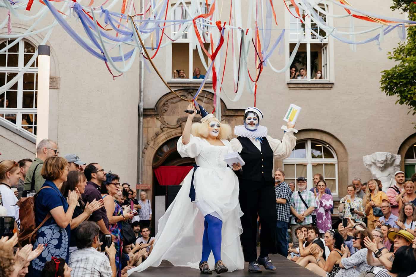 Ein belebter, heller Innenhof der HfBK Dresden während einer sommerlichen Modenschau vor einem historischen Gebäude.  Auf einem erhöhten Laufsteg stehen zwei Darsteller in barock-clownesken, schwarz-weißen Kostümen mit aufwendigem Gesichts-Make-up.