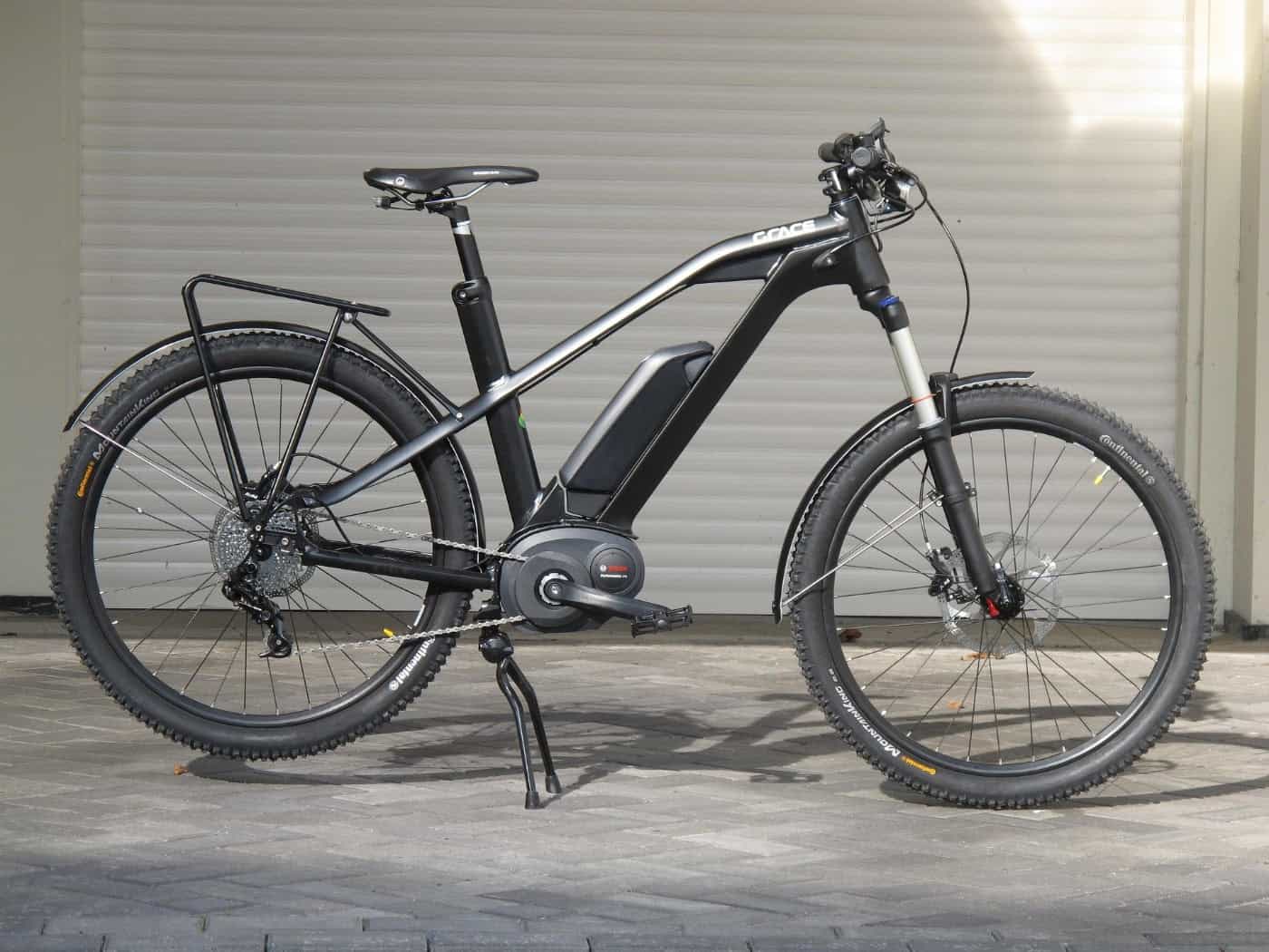 Ein sportliches, schwarzes E-Bike mit sichtbarem Rahmenakku, Mittelmotor und Packtaschenhalterung steht auf gepflastertem Boden vor einem weißen Rolltor.