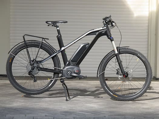 Ein sportliches, schwarzes E-Bike mit sichtbarem Rahmenakku, Mittelmotor und Packtaschenhalterung steht auf gepflastertem Boden vor einem weißen Rolltor.