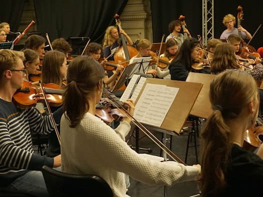 Ein Jugendorchester bei einer Probe. Im Vordergrund spielen Jugendliche konzentriert Violine, im Hintergrund sind Kontrabässe und Notenständer auf einer Bühne vor dunklem Vorhang zu sehen.