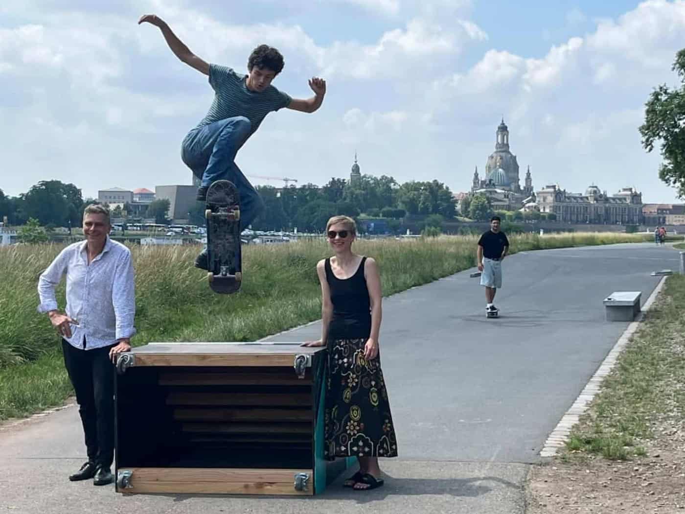 Drei Personen am asphaltierten Elberadweg vor der Silhouette der Dresdner Altstadt mit der Frauenkirche. Links steht ein Mann im hellen Hemd, mittig eine Frau im schwarzen Top und gemusterten Rock. Im Vordergrund springt ein junger Skater mit seinem Board hoch über eine mobile Holzrampe.