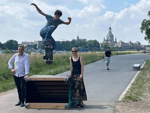 Drei Personen am asphaltierten Elberadweg vor der Silhouette der Dresdner Altstadt mit der Frauenkirche. Links steht ein Mann im hellen Hemd, mittig eine Frau im schwarzen Top und gemusterten Rock. Im Vordergrund springt ein junger Skater mit seinem Board hoch über eine mobile Holzrampe.