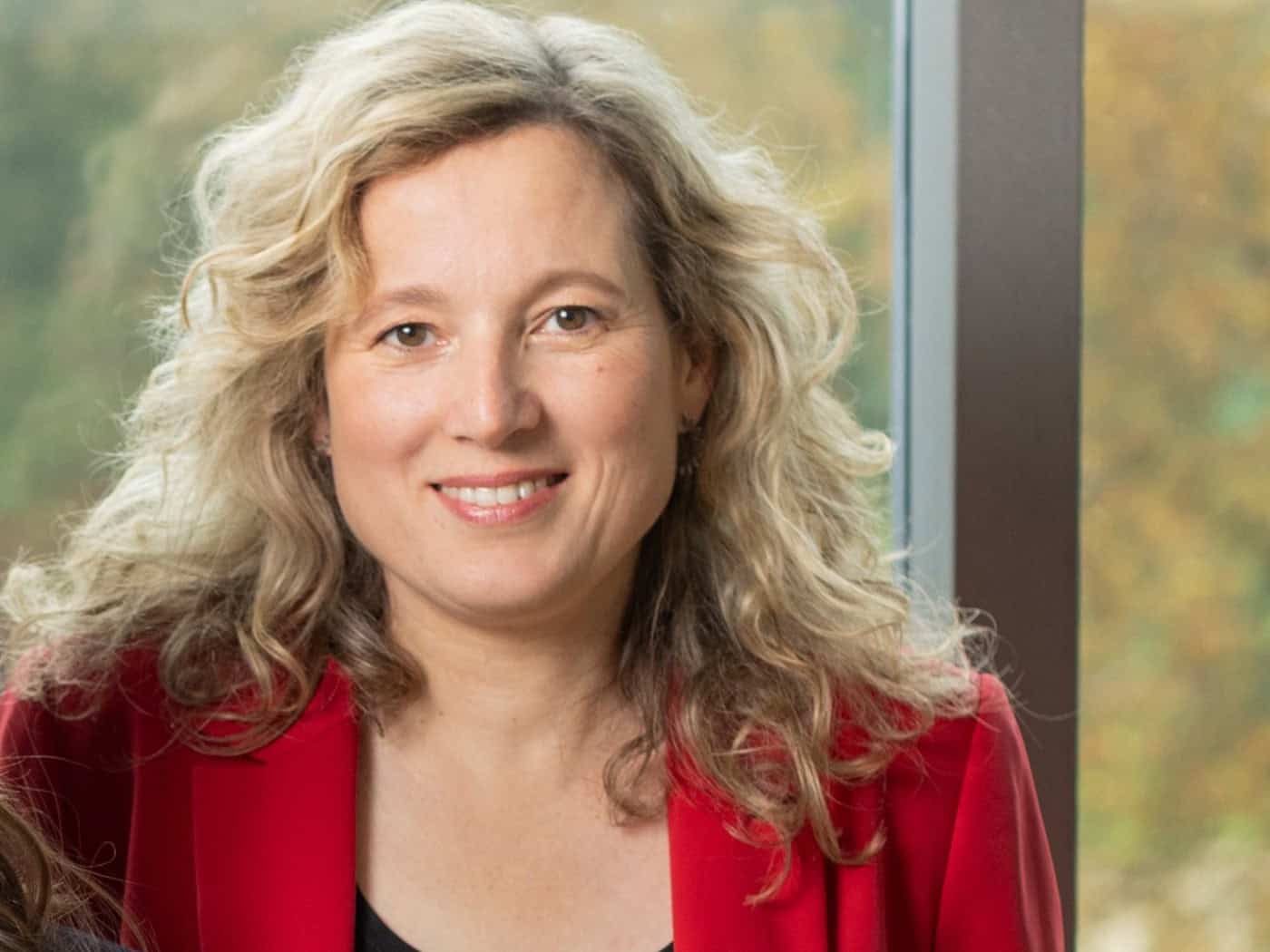 Porträt einer lächelnden Frau mit schulterlangen, blonden, lockigen Haaren. Sie trägt einen leuchtend roten Blazer über einem schwarzen Oberteil und blickt direkt in die Kamera vor einem hellen Hintergrund an einem Fenster.