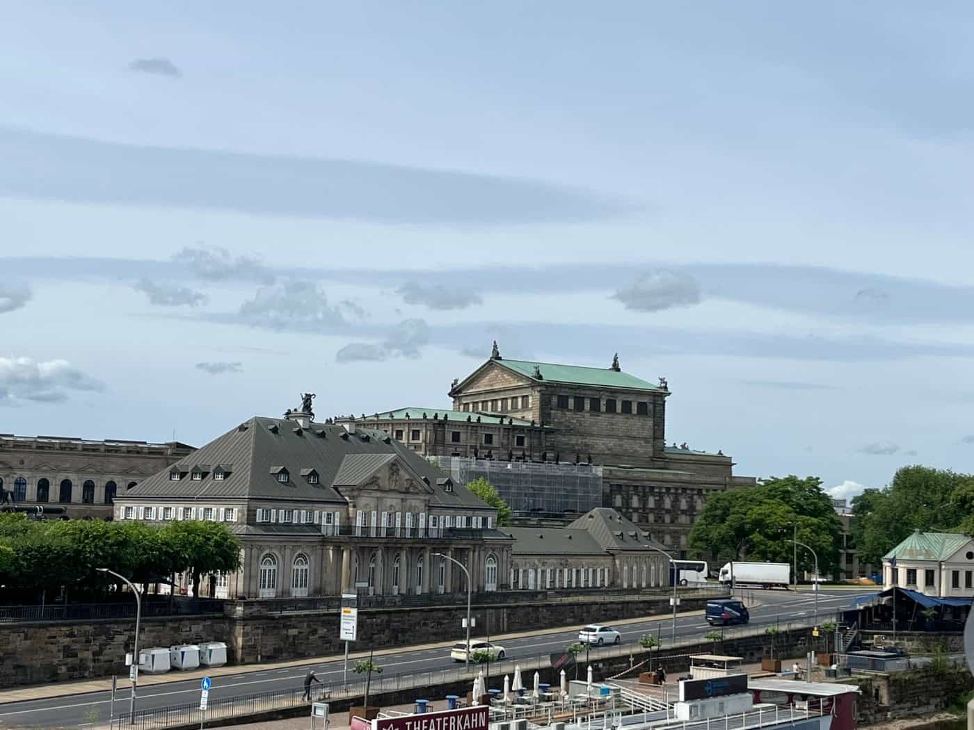 Eine Fotografie des historischen Gebäudes „Italienisches Dörfchen“ in Dresden, gelegen an einer Straße direkt am Elbufer. Dahinter erhebt sich die prachtvolle, sandsteinfarbene Fassade der Semperoper mit ihrem markanten grünen Kupferdach. Im Vordergrund am Flussufer ist das Dach des Theaterkahns zu 