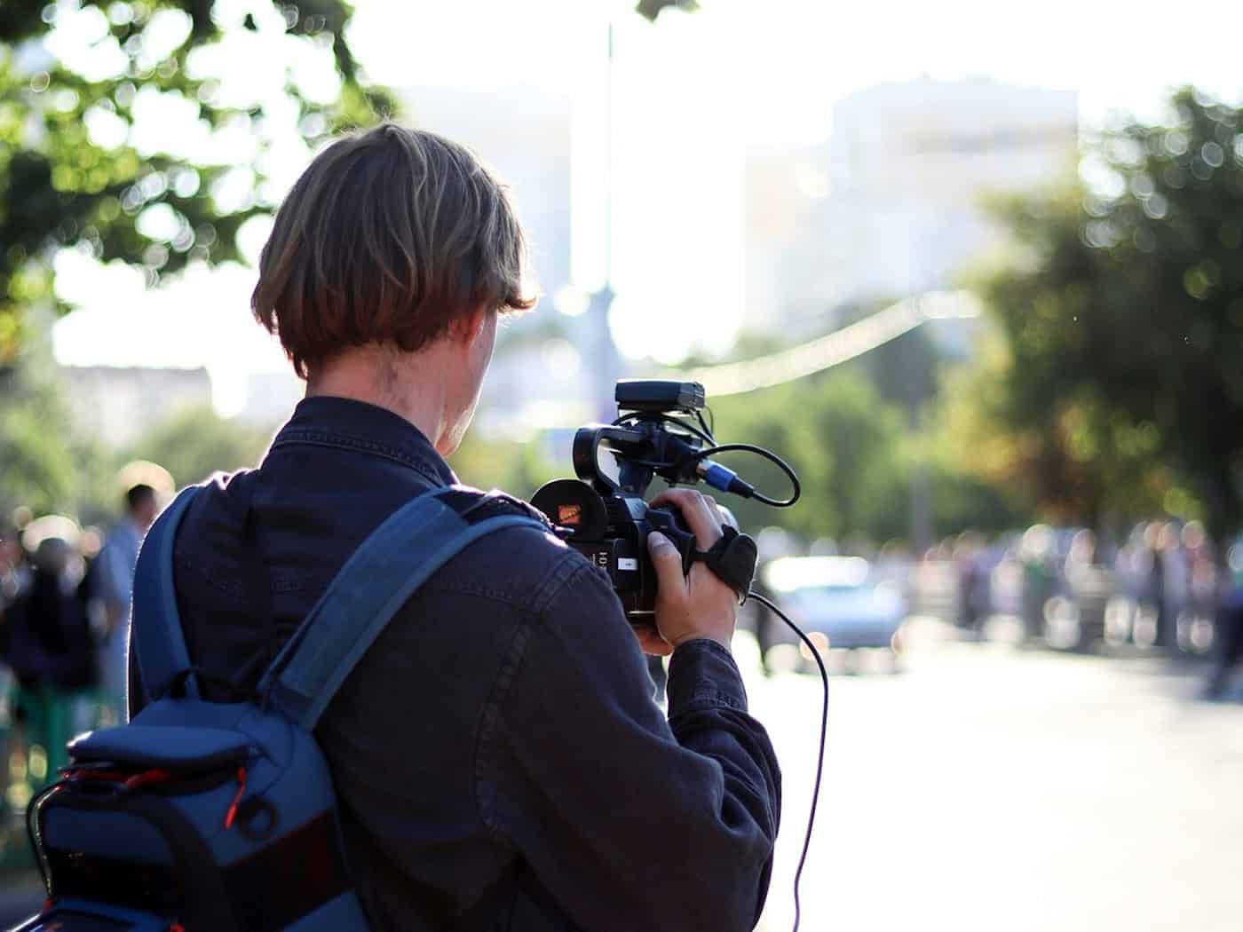 Foto: eine Person ist von Hinten zu sehen, mit einer professionellen Kamera etwas filmend, im Hintergrund nur unscharf Personen, Bäume und ein Auto