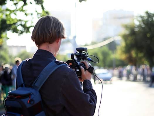 Foto: eine Person ist von Hinten zu sehen, mit einer professionellen Kamera etwas filmend, im Hintergrund nur unscharf Personen, Bäume und ein Auto
