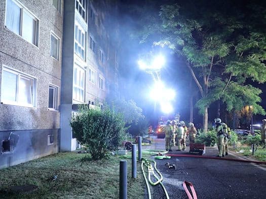 Nächtlicher Feuerwehreinsatz an einem Plattenbau. Ein ausgefahrener Leiterkorb beleuchtet die verrauchte Fassade mit Scheinwerfern. Im Vordergrund stehen Einsatzkräfte neben Schläuchen.