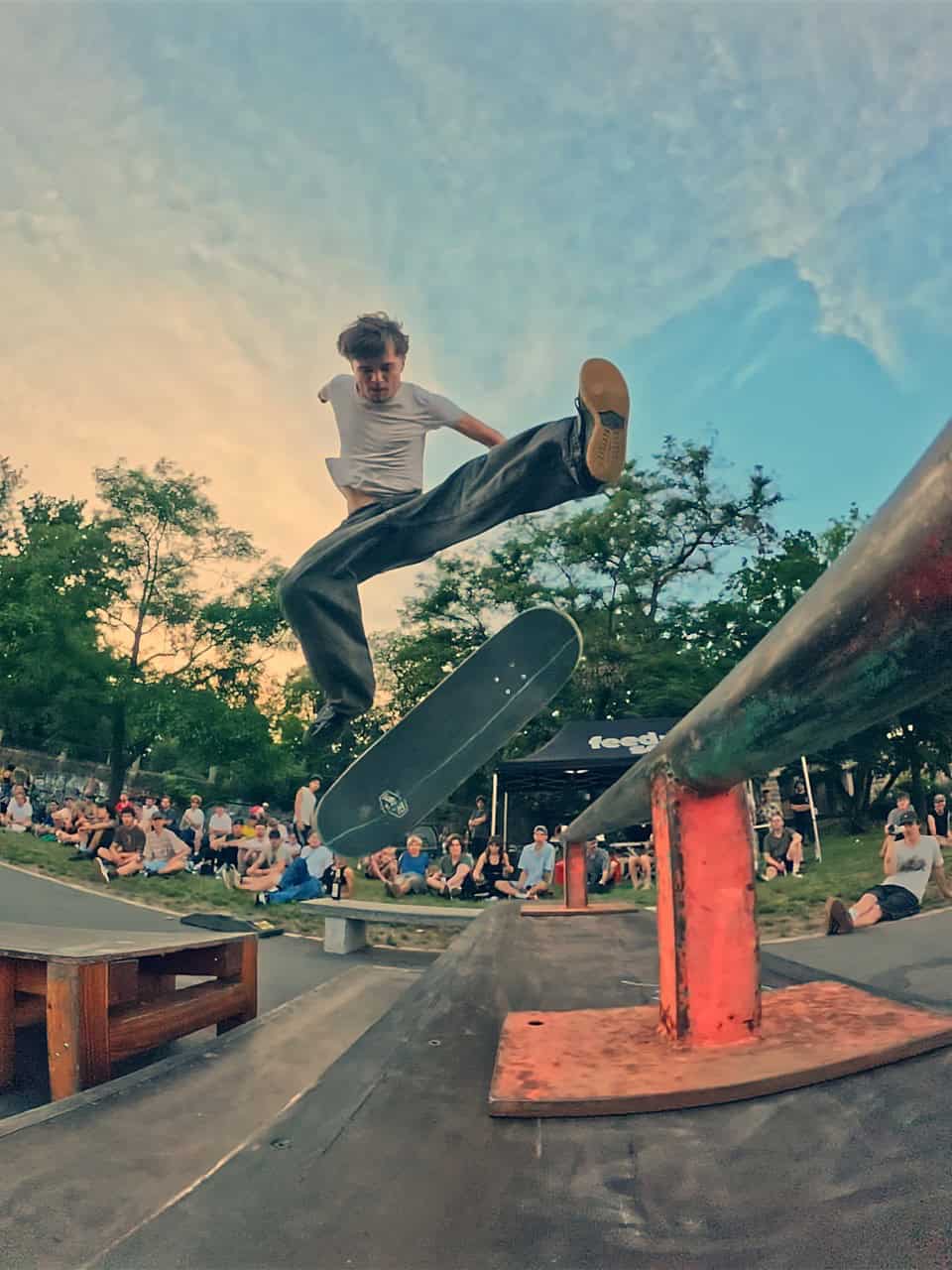 Dynamische Nahaufnahme aus einer tiefen Perspektive. Ein Skater in weißem T-Shirt und weiter Hose springt mit seinem Skateboard über eine rot-grüne Metallstange (Rail). Im Hintergrund sitzen Zuschauer auf einer Wiese unter Bäumen.