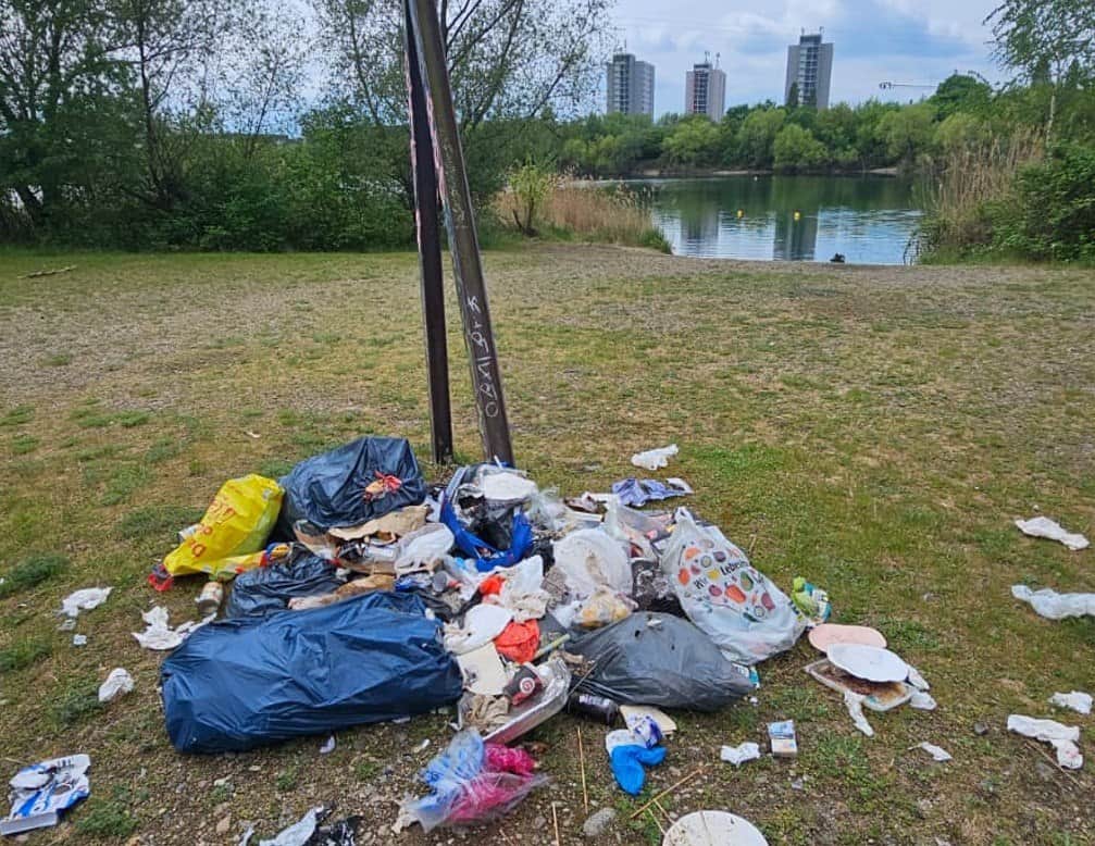 Ein großer Haufen wilder Müll mit blauen Plastiksäcken, weggeworfenen Papptellern und Verpackungen liegt auf einer Wiese am Ufer des Kiessees Leuben. Im Hintergrund sind auf der anderen Seeseite drei Plattenbauten zu sehen.