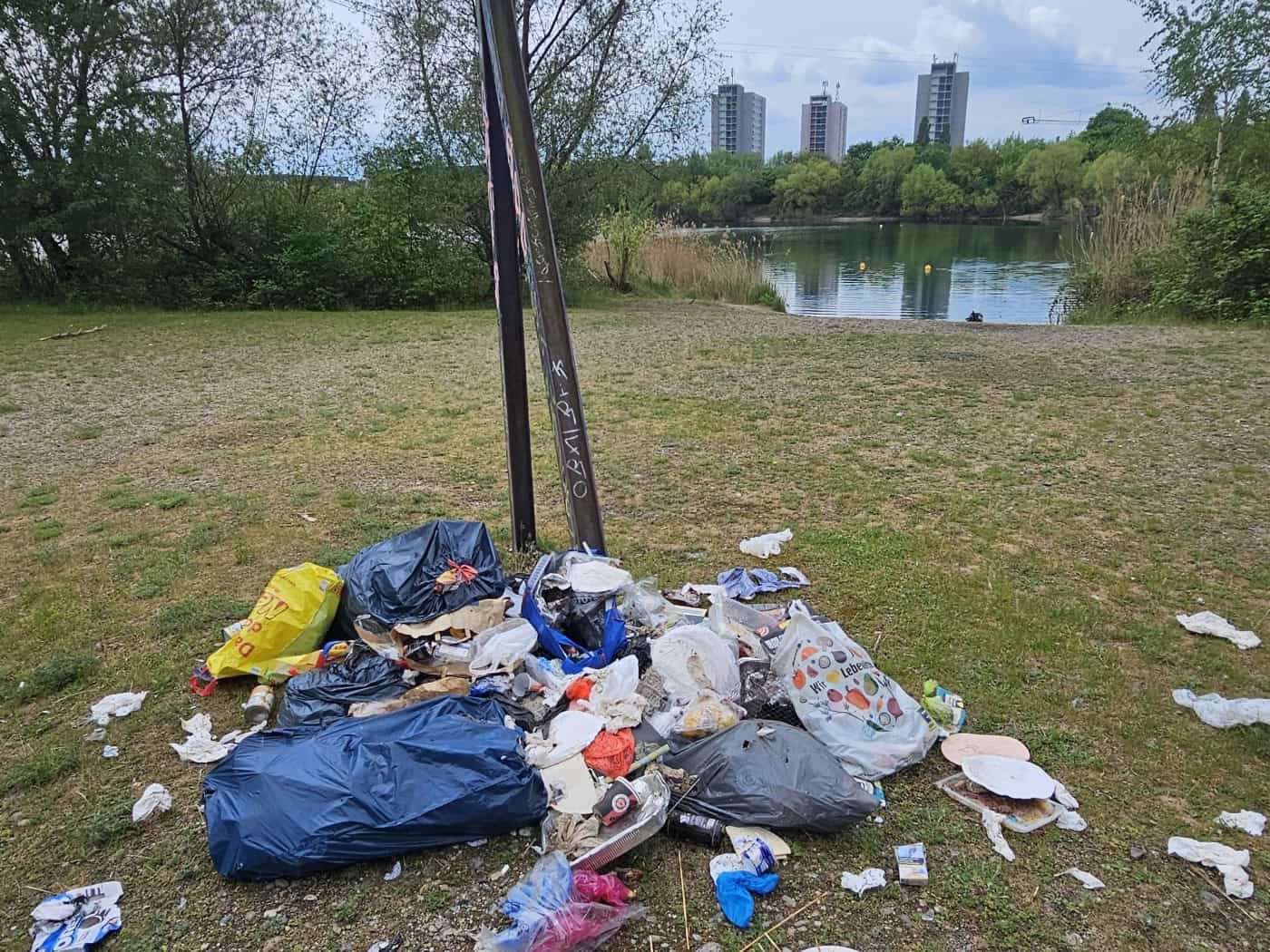 Ein großer Haufen wilder Müll aus Plastiktüten, Papptellern und Dosen auf einer Wiese. Im Hintergrund sieht man einen See, grüne Bäume und drei weit entfernte Wohnhochhäuser unter bewölktem Himmel.